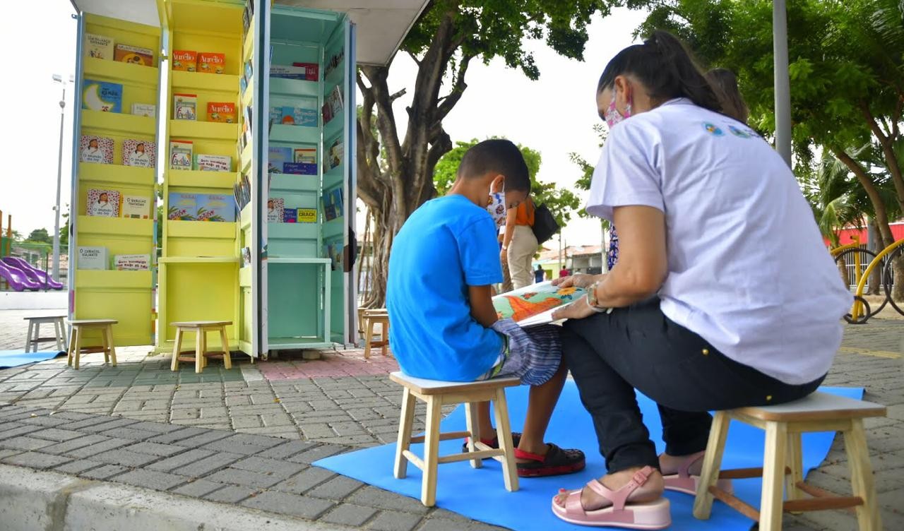 uma mulher e uma criança leem um livro na frente de um quiosque do projeto leitura na praça